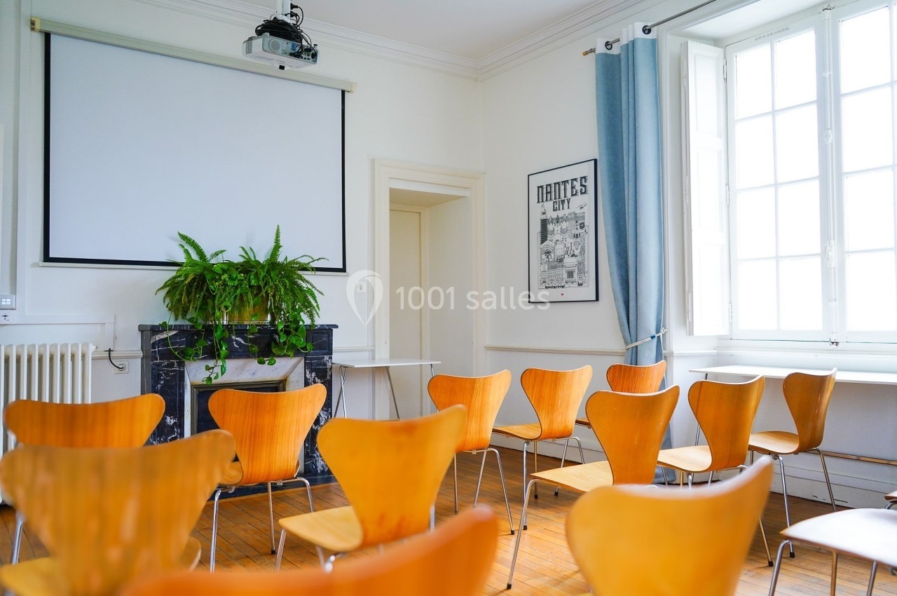 Salle de réunion lumineuse avec des chaises en bois, un projecteur, un écran et une plante sur une cheminée.