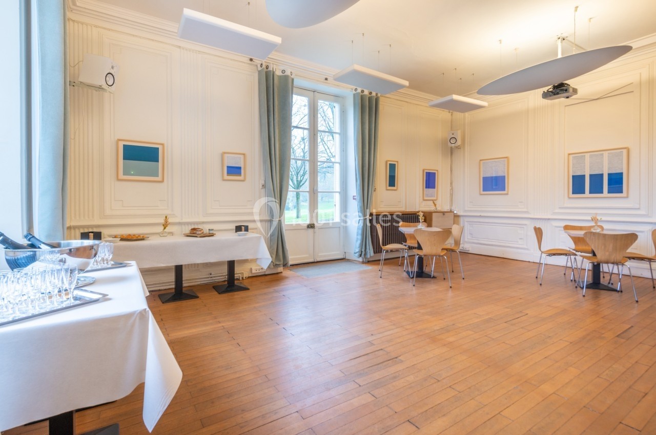Salle lumineuse avec parquet, tables et chaises en bois, murs ornés de cadres, et buffet dressé près des fenêtres.