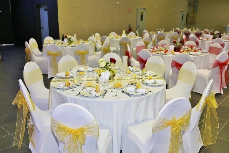 Salle de réception décorée avec des tables rondes, nappes blanches, chaises ornées de nœuds rouges et jaunes.