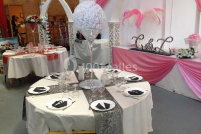 Salle décorée pour un événement, avec des tables rondes dressées, chaises rouges et un fond floral rose.