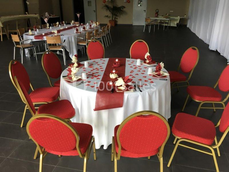 Salle décorée pour un événement, table ronde avec nappes blanches, chaises rouges et décorations rouges et blanches.