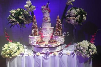 Salle décorée pour un événement, avec des tables rondes dressées, chaises rouges et un fond floral rose.