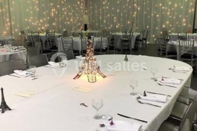 Salle décorée pour un événement, avec des tables rondes dressées, chaises rouges et un fond floral rose.