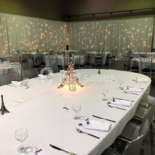 Salle de réception décorée avec des tables dressées, des chaises argentées et des guirlandes lumineuses en arrière-plan.