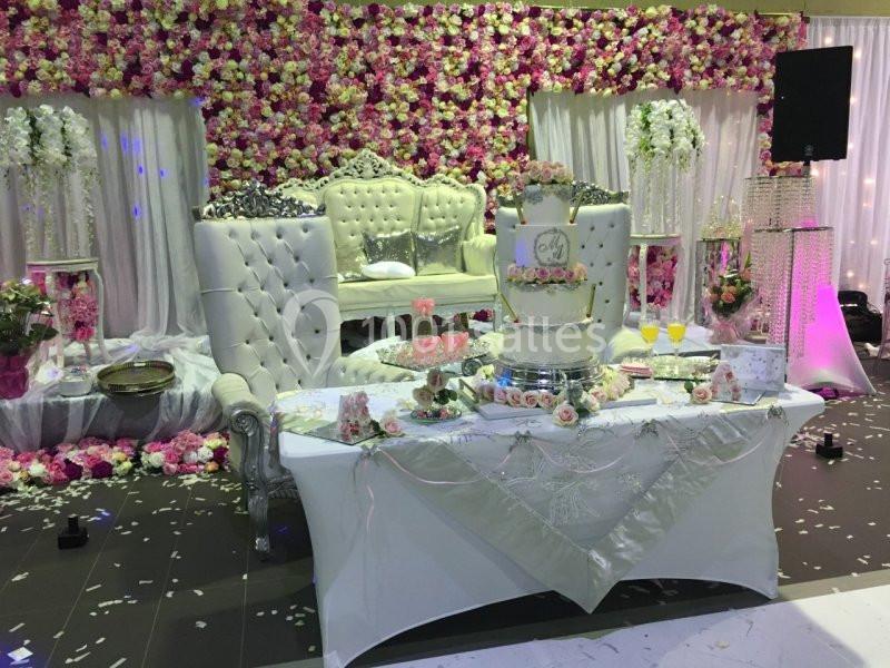 Décoration de mariage avec table ornée d'un gâteau à étages, fauteuils blancs capitonnés et mur de fleurs colorées.