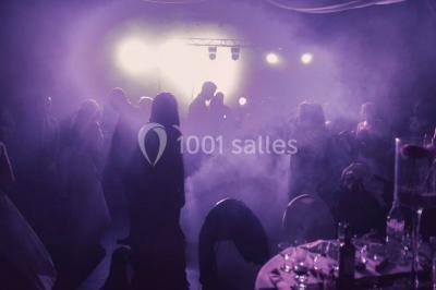 Salle décorée pour un événement, avec des tables rondes dressées, chaises rouges et un fond floral rose.