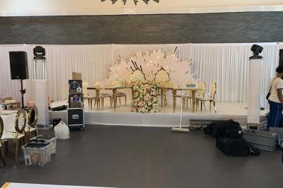 Salle décorée pour un événement, avec des tables rondes dressées, chaises rouges et un fond floral rose.