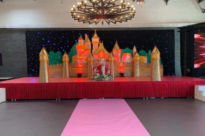 Salle décorée pour un événement, avec des tables rondes dressées, chaises rouges et un fond floral rose.