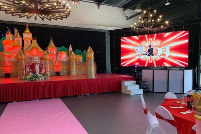 Salle décorée pour un événement, avec des tables rondes dressées, chaises rouges et un fond floral rose.