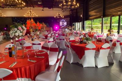 Salle décorée pour un événement, avec des tables rondes dressées, chaises rouges et un fond floral rose.