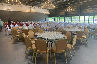 Salle décorée pour un événement, avec des tables rondes dressées, chaises rouges et un fond floral rose.