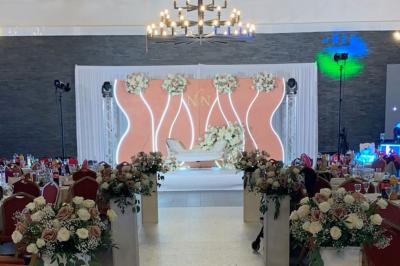 Salle décorée pour un événement, avec des tables rondes dressées, chaises rouges et un fond floral rose.