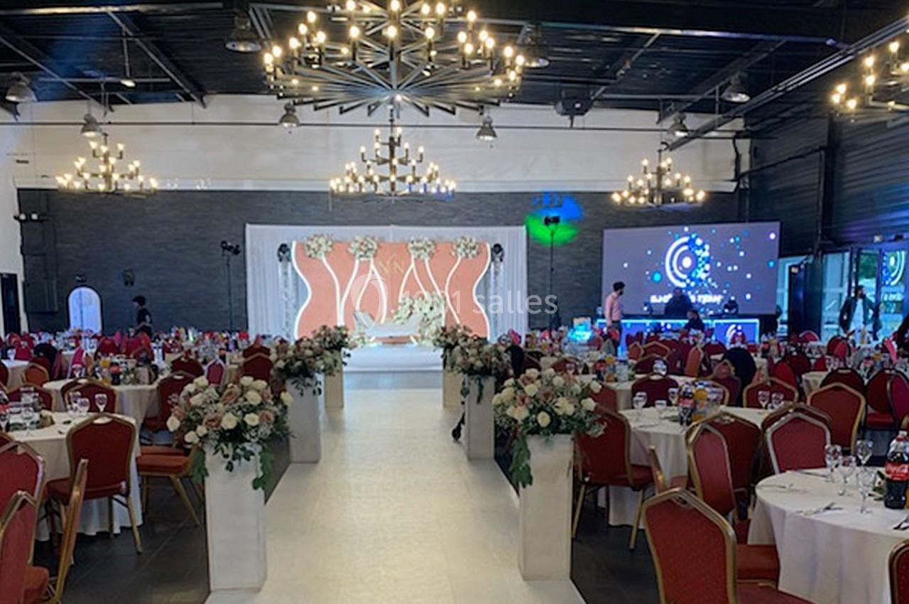 Salle de réception décorée avec des chaises rouges, des tables fleuries et une allée centrale blanche menant à une scène.