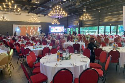 Salle décorée pour un événement, avec des tables rondes dressées, chaises rouges et un fond floral rose.