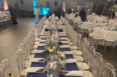 Salle décorée pour un événement, avec des tables rondes dressées, chaises rouges et un fond floral rose.