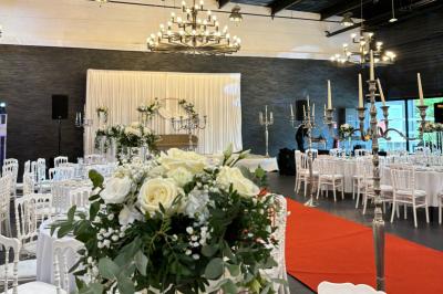 Salle décorée pour un événement, avec des tables rondes dressées, chaises rouges et un fond floral rose.