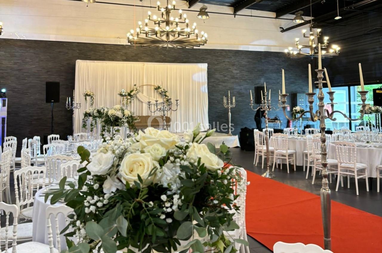 Salle de réception décorée avec des chandeliers, des fleurs blanches et un tapis rouge menant à une scène ornée de rideaux…