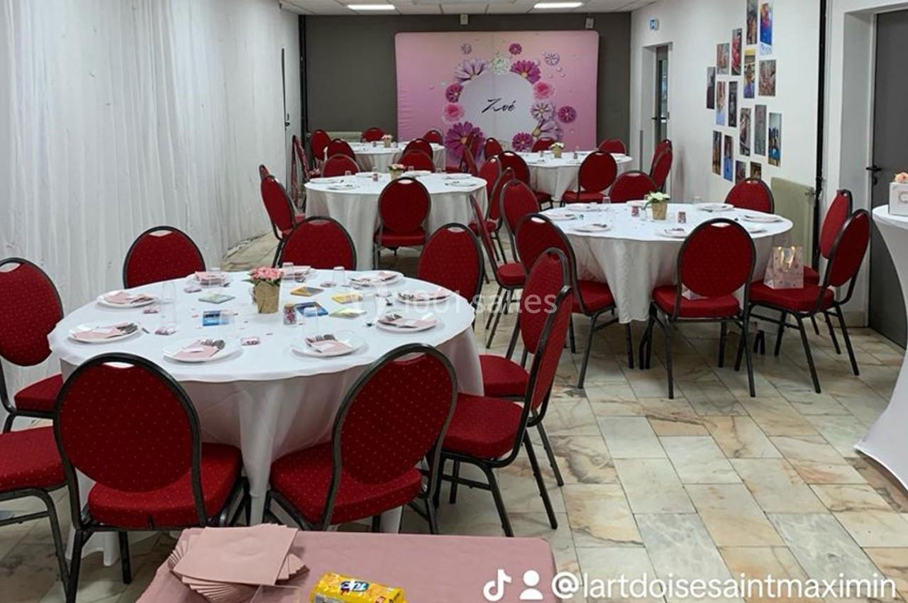Salle décorée pour un événement, avec des tables rondes dressées, chaises rouges et un fond floral rose.