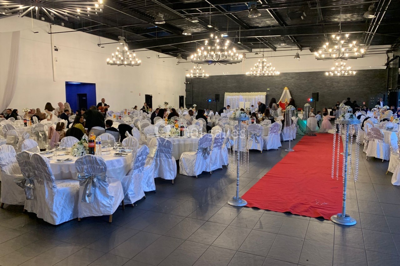Salle de réception décorée avec des tables dressées, des chaises blanches et un tapis rouge menant à une scène.