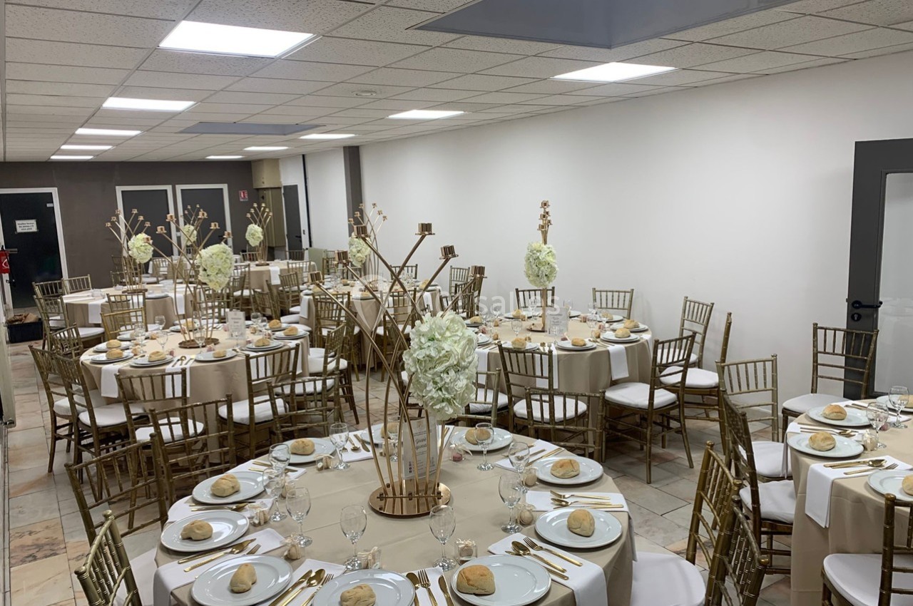 Salle de réception décorée avec des tables rondes dressées, chaises dorées et centres de table floraux.