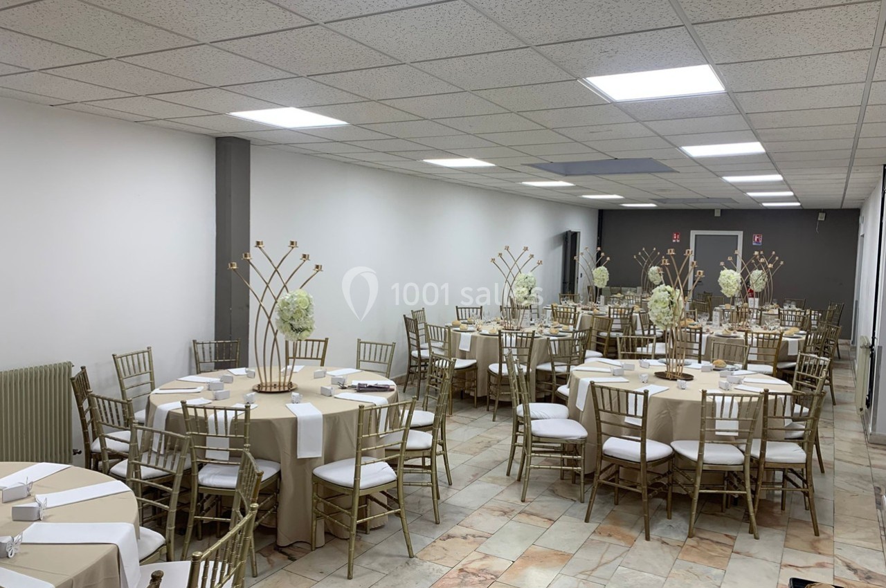 Salle de réception décorée avec des tables rondes, nappes beige, chaises dorées et centres de table floraux.