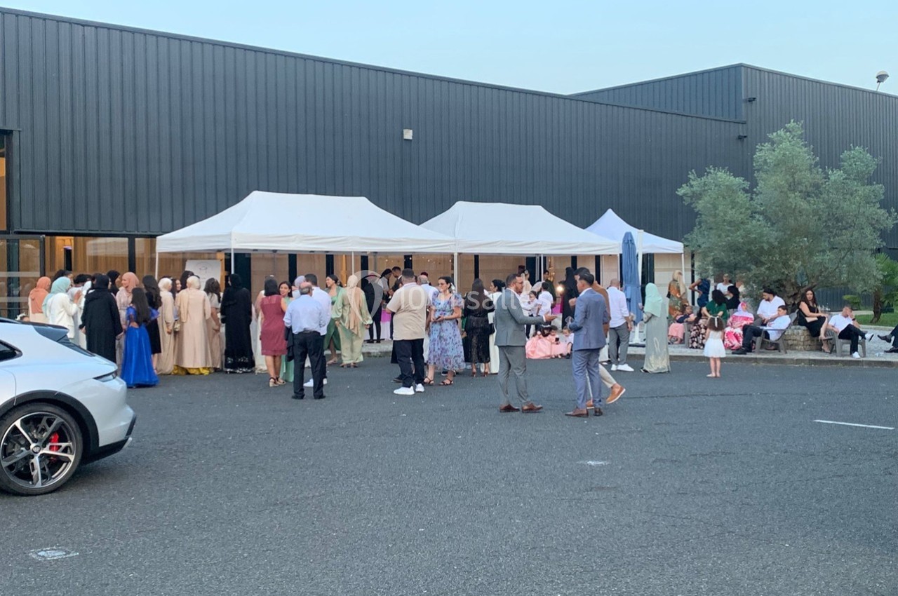 Groupe de personnes rassemblées devant des tentes blanches sur un parking, près d'un bâtiment moderne.