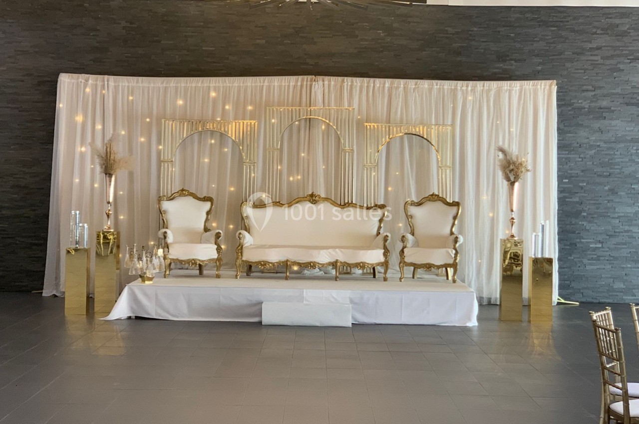 Décor de cérémonie avec trois fauteuils dorés, rideau blanc éclairé et colonnes dorées ornées de pampas.