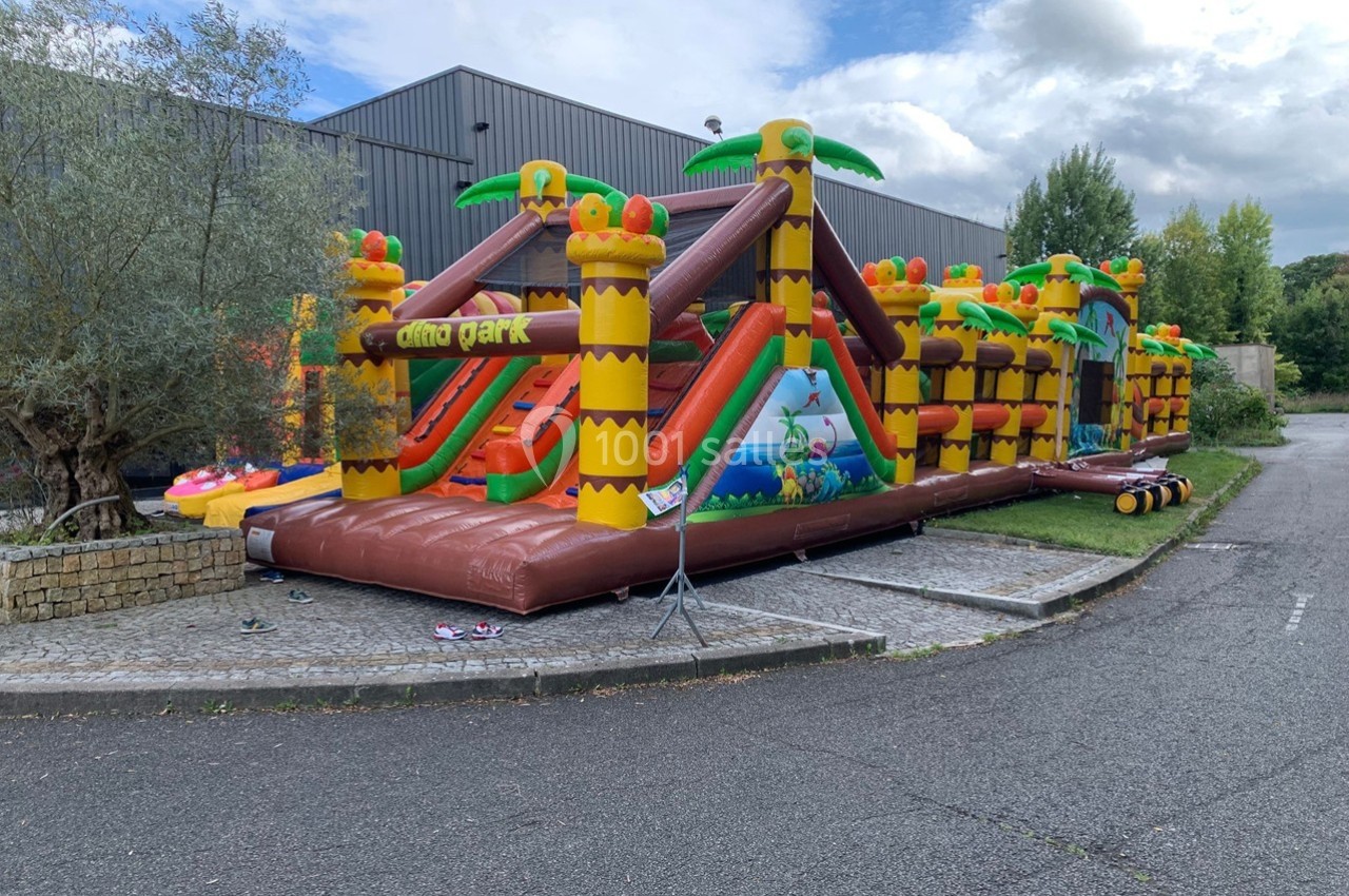 Structure gonflable colorée sur le thème des dinosaures avec toboggans, installée en extérieur près d'un bâtiment.