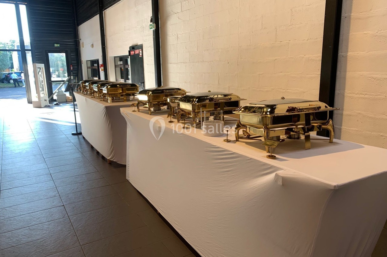 Buffet avec plusieurs plats en inox sur des tables recouvertes de nappes blanches dans une salle lumineuse.