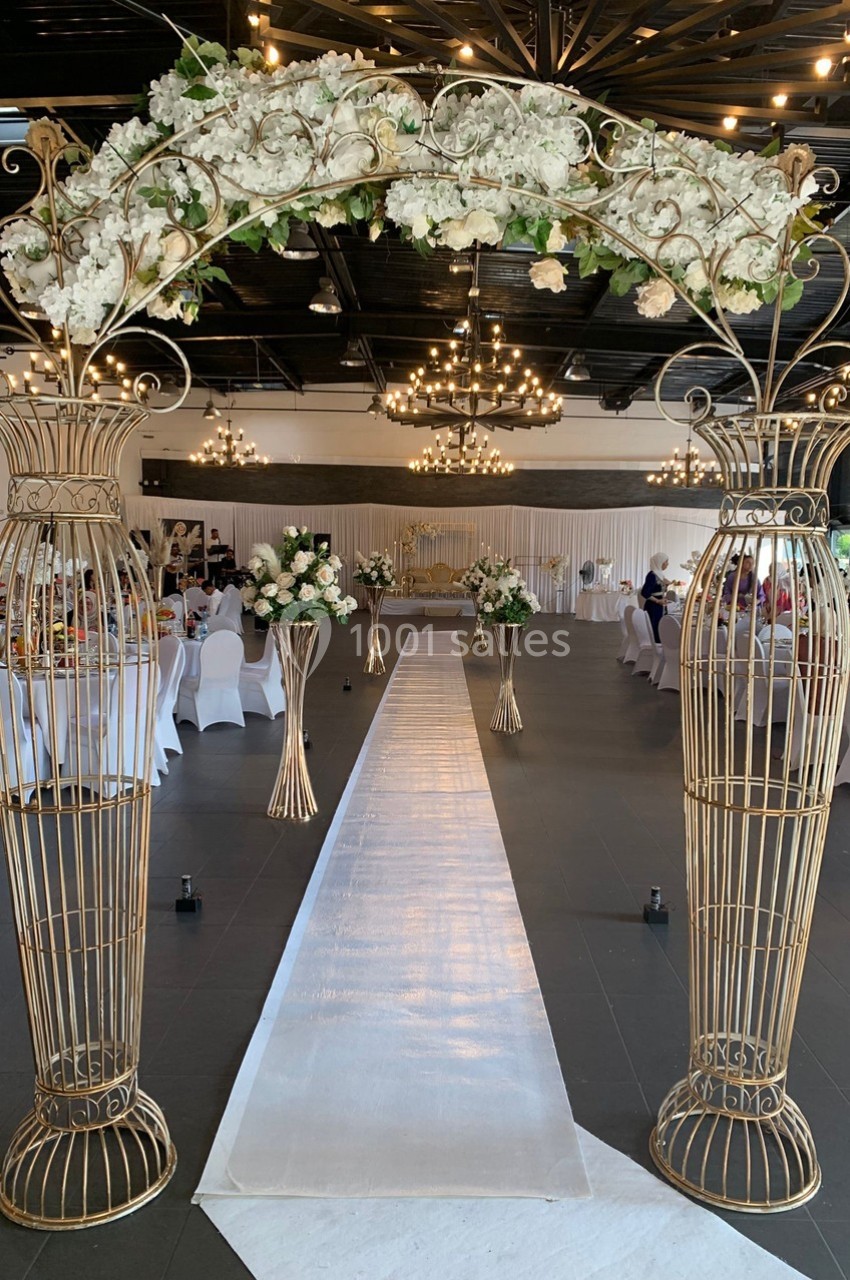 Allée de mariage décorée avec un tapis blanc, des arches dorées ornées de fleurs blanches et des chaises drapées.