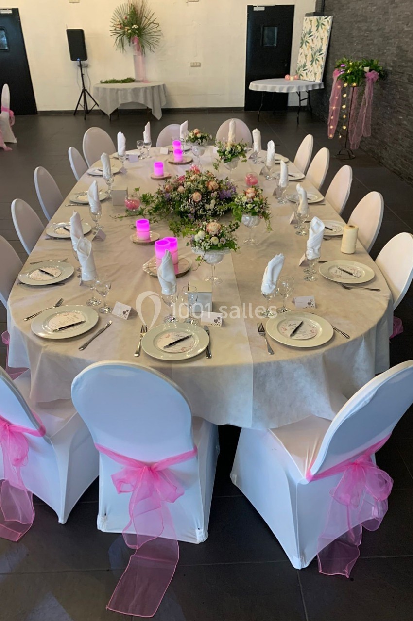 Table ovale décorée pour un événement, avec nappes blanches, chaises ornées de rubans roses et centre de table floral.