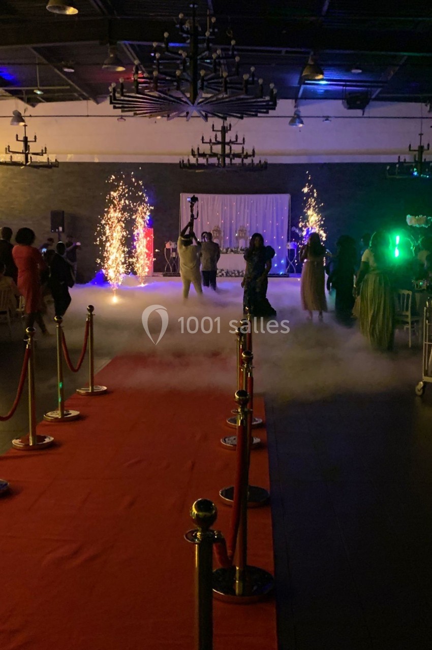 Salle de réception avec tapis rouge, invités dansant sous des effets de fumée et étincelles lumineuses.