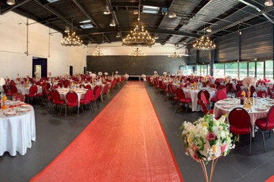 Salle décorée avec des tables rondes, nappes blanches et chaises rouges, prête pour un événement festif.