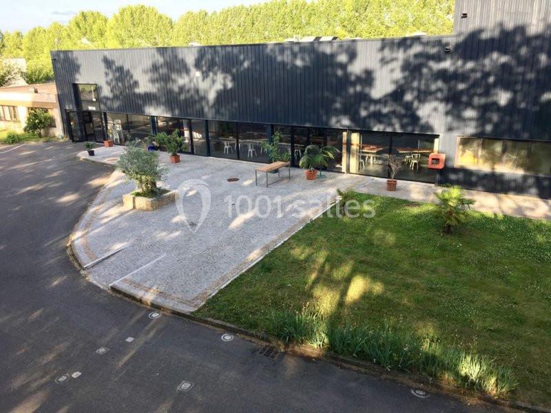 Bâtiment moderne avec façade sombre, entouré d'un espace vert, d'une terrasse pavée et de plantes en pots.