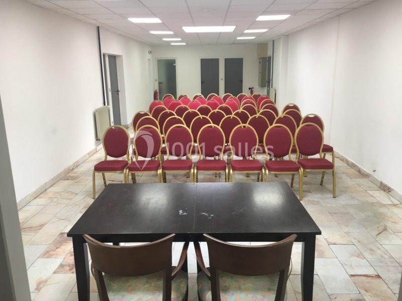 Salle de réunion avec des rangées de chaises rouges alignées face à une table noire et deux chaises.
