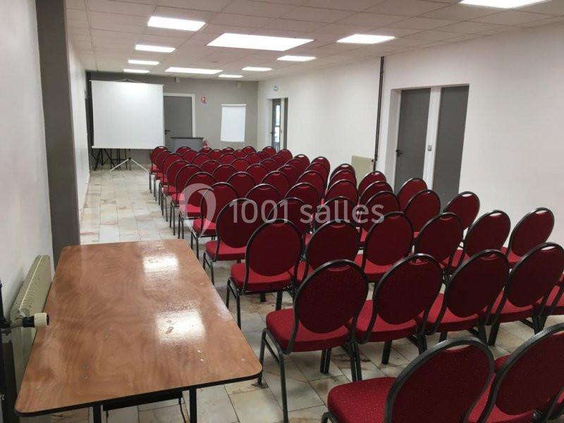 Salle de conférence avec des rangées de chaises rouges alignées, un écran de projection et un tableau blanc.