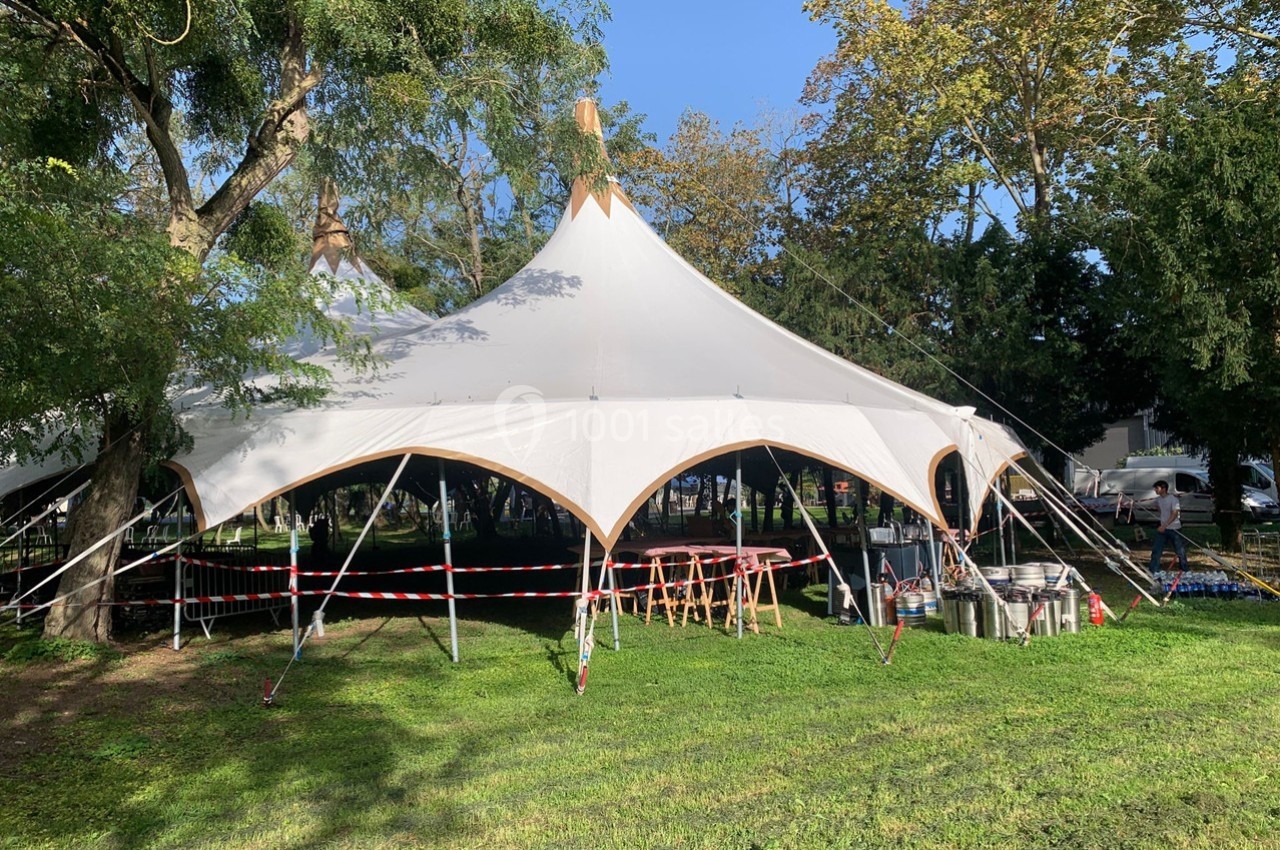 Grande tente blanche installée dans un parc verdoyant, entourée d'arbres et de matériel d'aménagement.