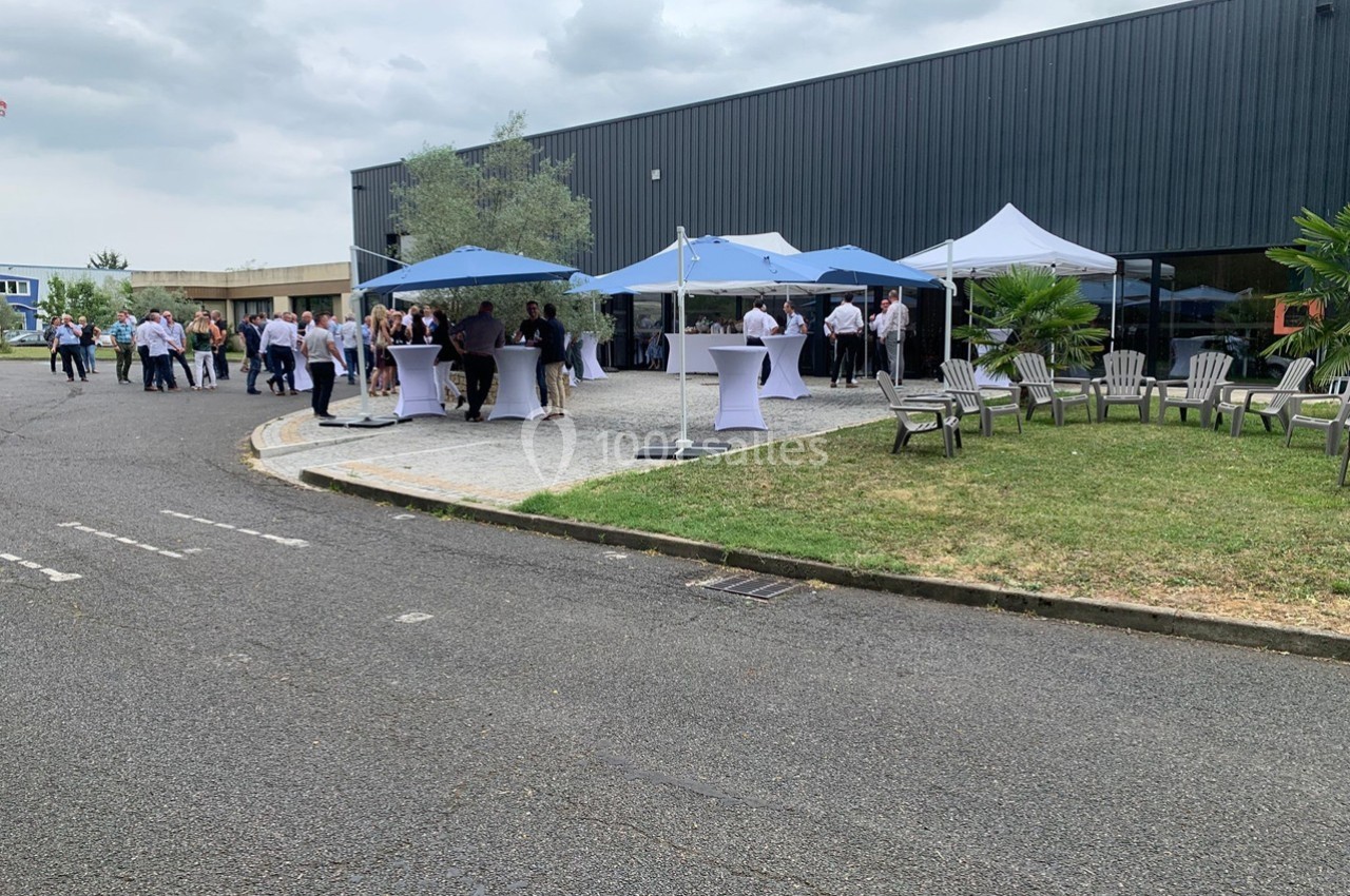 Personnes rassemblées autour de tables sous des tentes blanches et bleues dans une cour extérieure près d'un bâtiment.