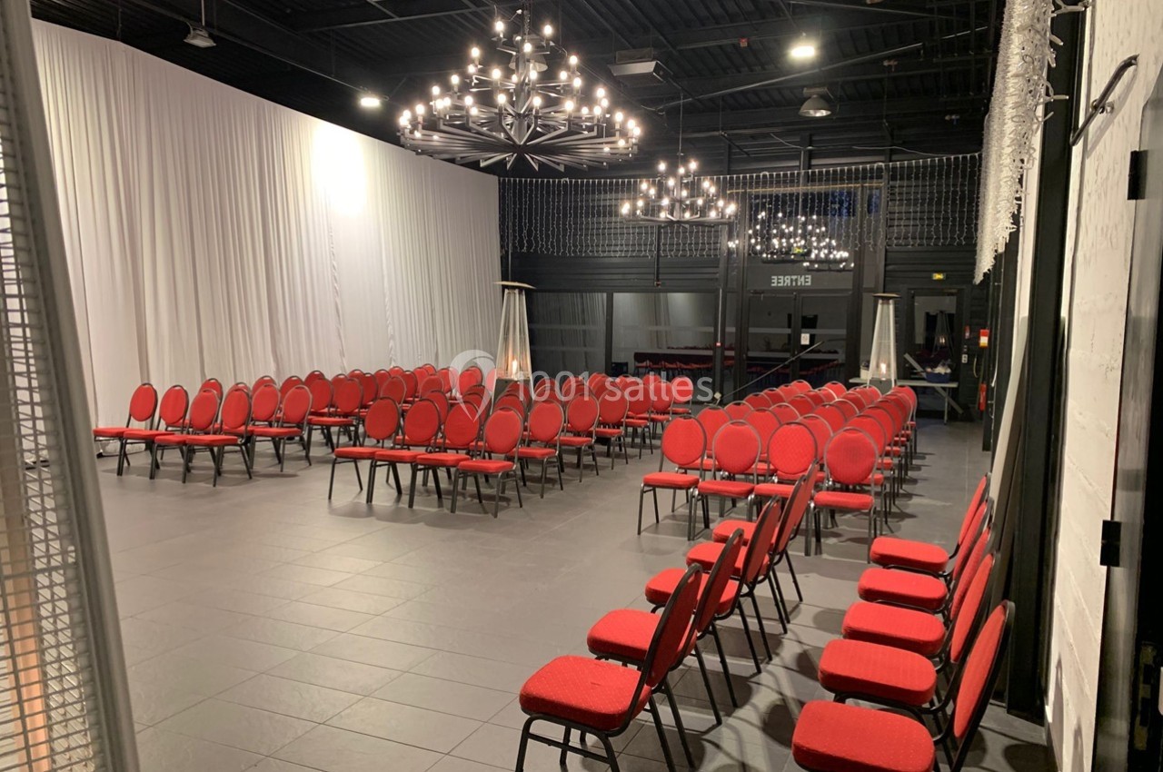 Salle de réception avec des rangées de chaises rouges alignées sous des lustres modernes dans un espace sombre.