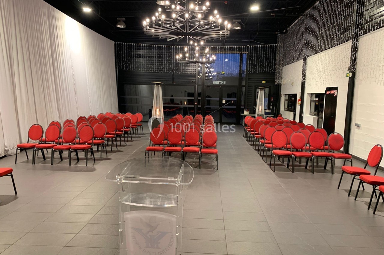 Salle de conférence avec des rangées de chaises rouges alignées face à un pupitre transparent, éclairée par des lustres…