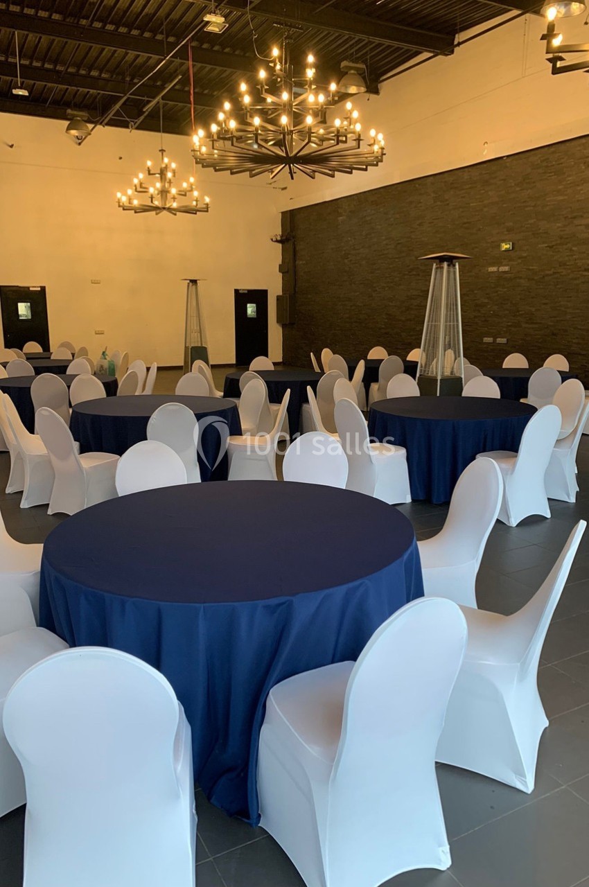 Salle de réception avec tables rondes nappées de bleu, chaises blanches et lustres suspendus au plafond.