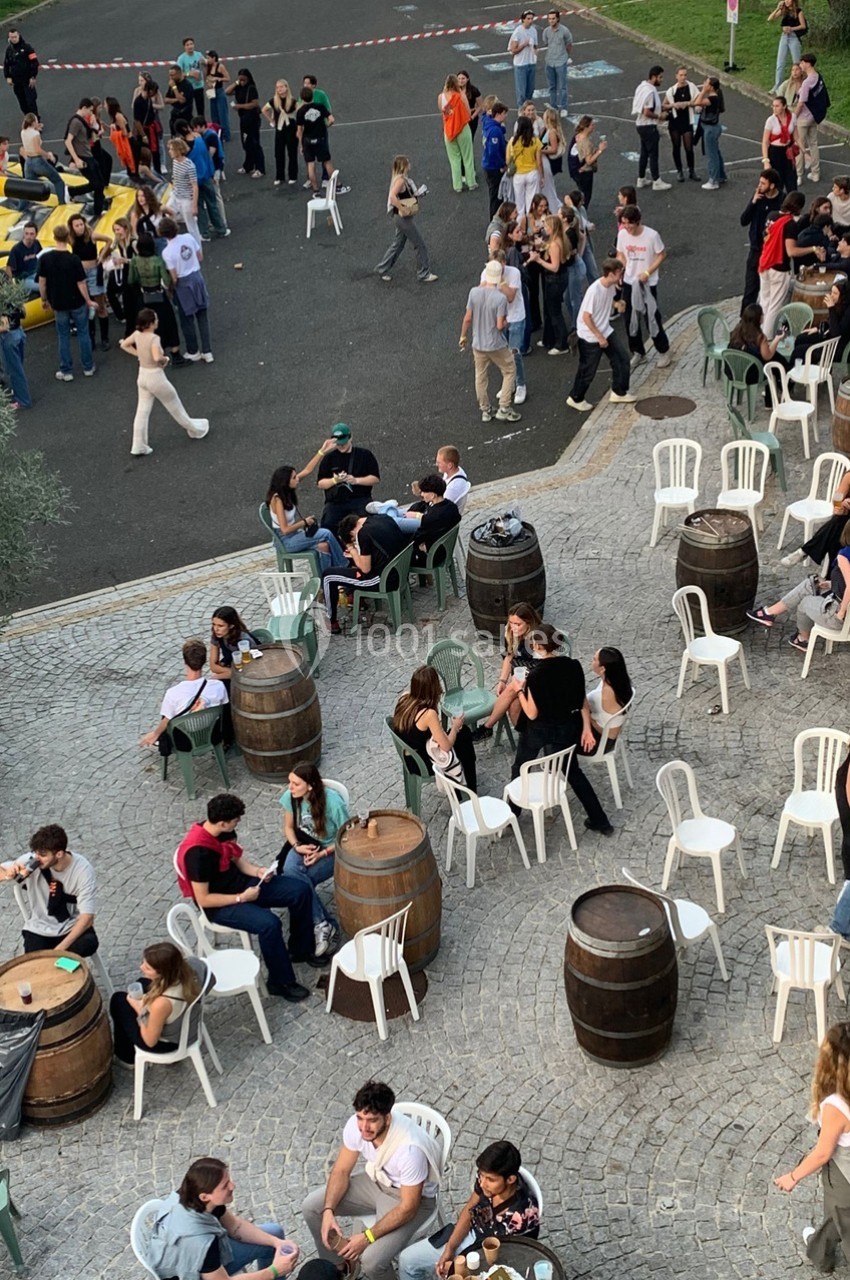 Groupe de personnes assises et debout lors d'un événement en plein air, entourées de tables et tonneaux sur une place pavée.