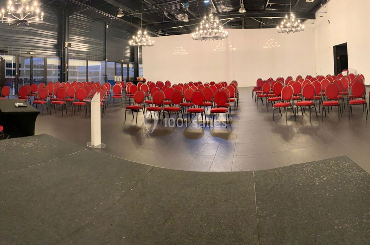 Salle de conférence vide avec des rangées de chaises rouges alignées face à une estrade et un pupitre blanc.