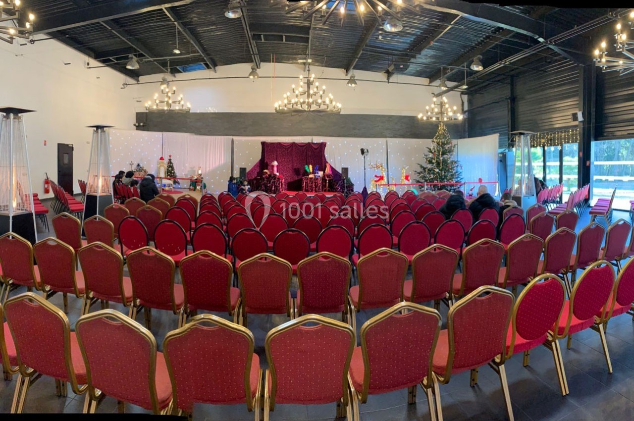 Salle de réception avec des rangées de chaises rouges, une scène décorée et un sapin de Noël à droite.