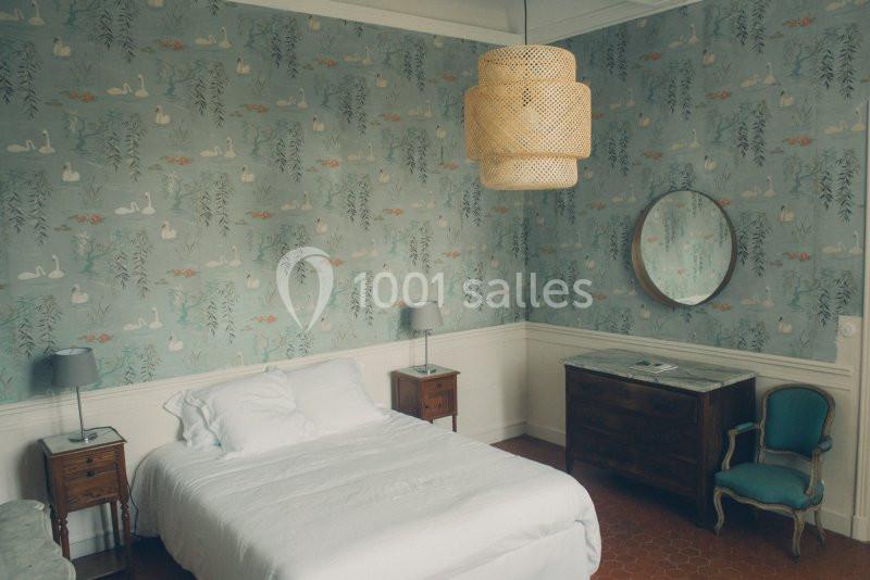 Chambre lumineuse avec lit double, murs ornés de papier peint à motifs de cygnes, mobilier en bois et fauteuil ancien.