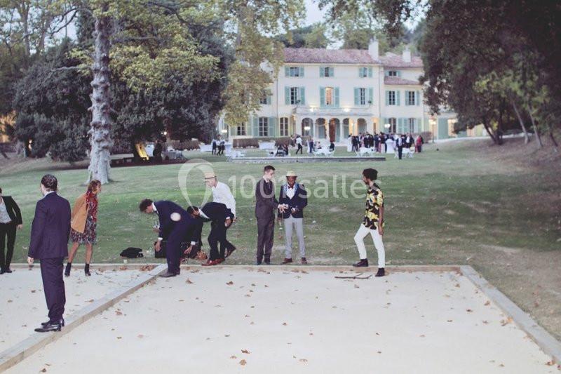 Des personnes jouent à la pétanque sur un terrain devant une grande maison entourée d'arbres.