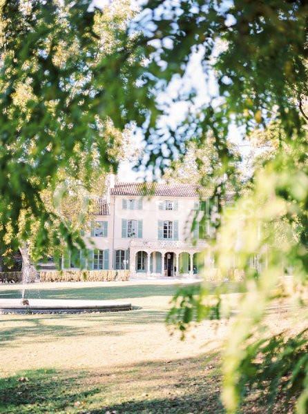 Grande maison provençale entourée d'arbres, vue à travers un feuillage, sous une lumière douce.