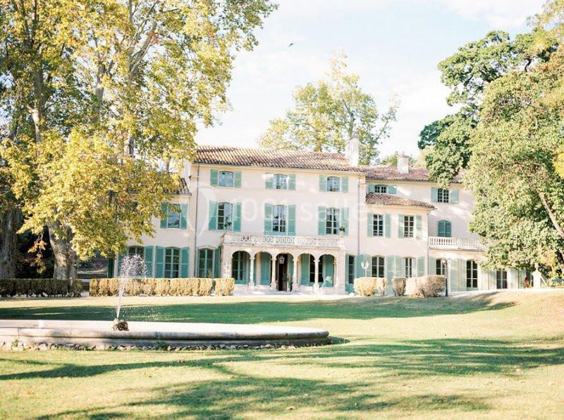 Grande maison provençale entourée d'arbres, avec une pelouse et une fontaine au premier plan.