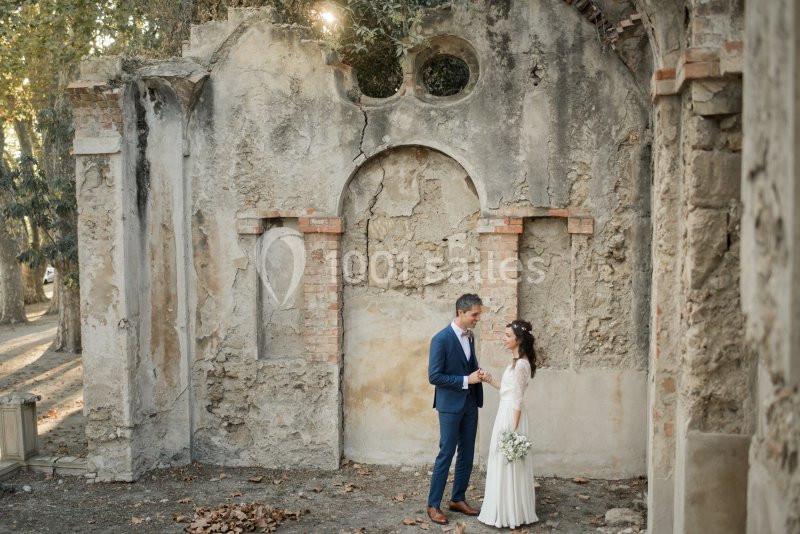 Un couple en tenue de mariage se tient devant un mur en pierre ancienne dans un cadre extérieur ombragé.