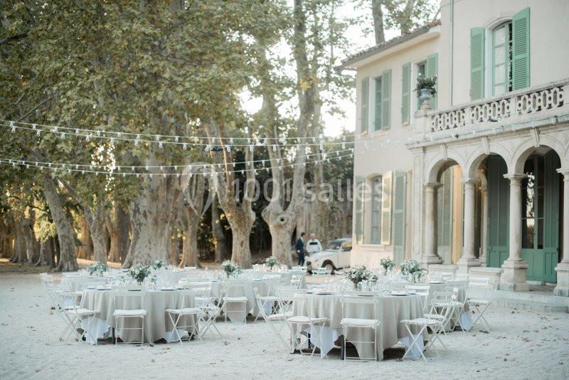 Tables rondes décorées pour un événement en plein air, disposées devant une maison avec volets verts et des arbres en…